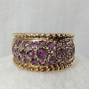 May include: A gold ring with a band of small, round, purple gemstones. The ring has a textured, beaded design on the top and bottom edges. The gemstones are set closely together, creating a continuous band of color.