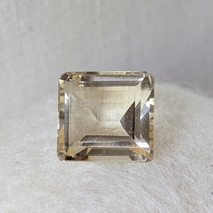 May include: A square-cut, translucent gemstone with a pale yellow, champagne-like hue. The stone features a faceted surface with sharp, geometric edges and a clear, reflective interior that displays light refraction through its transparent body.