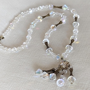 May include: A vintage crystal necklace with a dangling crystal tassel. The necklace is made of clear faceted crystal beads and has a silver clasp. The tassel is made of several smaller crystal beads and has a silver accent.