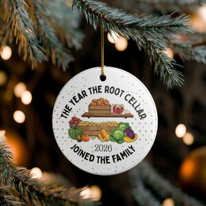 May include: A round white ceramic ornament featuring a gold hanging string. The design displays wooden crates filled with vegetables, jars, and the text "THE YEAR THE ROOT CELLAR JOINED THE FAMILY" along with the year "2026" in black lettering.