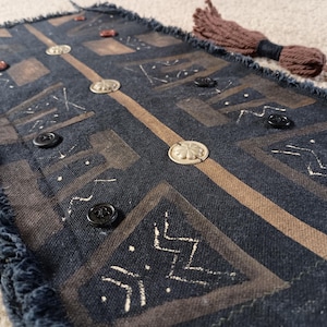 Puede incluir: Textil rectangular con borde deshilachado, con fondo azul oscuro con patrones geométricos marrones y detalles blancos. Botones plateados y negros dispersos por la superficie, junto con una borla marrón.