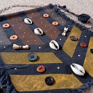 Puede incluir: Bolso textil cuadrado y con flecos con una cadena. El bolso presenta un fondo azul marino con patrones geométricos marrones y amarillos. Adornado con conchas, botones y cuentas en forma de hueso. El bolso tiene una borla azul marino.