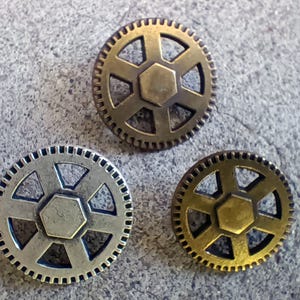 May include: Three bronze-colored gear-shaped buttons with a central hexagonal design and surrounding teeth. The buttons vary slightly in color, with one appearing silver, one gold, and one a darker bronze. The buttons are set against a textured gray surface.
