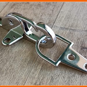 Könnte beinhalten: Ein silberner Metallhaken- und Schließverschluss mit einem D-Ring. Der Haken ist an einer Metallplatte mit zwei Löchern befestigt. Die Schlaufe ist an einer Metallplatte mit einem Loch befestigt.