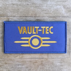 Könnte beinhalten: Ein blaues, rechteckiges Abzeichen mit dem goldenen Schriftzug "VAULT-TEC". Darunter befindet sich ein goldenes Emblem mit einem Kreis und drei horizontalen Linien auf jeder Seite. Der Aufnäher ist wahrscheinlich ein gestickter Artikel.