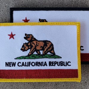 Op de afbeelding: Een met geel omzoomde patch met een witte achtergrond, met een rode ster, twee bruine beren en de tekst "NEW CALIFORNIA REPUBLIC". De patch heeft een rode streep aan de onderkant.