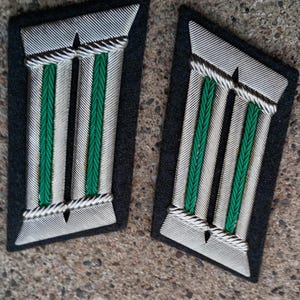 May include: A pair of rectangular dark grey collar tabs featuring two vertical silver metallic bars separated by a central green braided stripe. Each tab has a silver braided border at the top and bottom, creating a structured, symmetrical design.