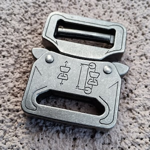 May include: A pair of silver-colored metal buckles, one on top of the other. The top buckle has a rectangular shape with a bar. The bottom buckle has a more complex design with a series of lines and symbols etched into the surface.