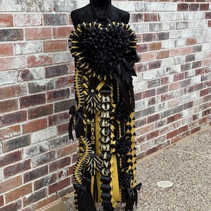 Puede incluir: Un accesorio de homecoming mum en negro y dorado con un gran centro floral en forma de corazón en la parte superior, cintas en cascada y lazos decorativos. La cinta vertical muestra la palabra HOMECOMING y JUNIOR en letras doradas.
