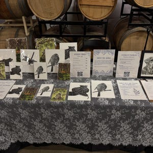 May include: A display table covered with a grey lace tablecloth featuring various framed and unframed wildlife art prints, informational signs with QR codes, and a clipboard with a pen. The prints depict birds, animals, and pet portraits.