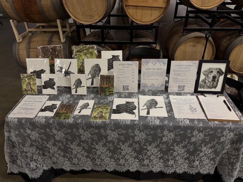 May include: A display table covered with a grey lace tablecloth featuring various framed and unframed wildlife art prints, informational signs with QR codes, and a clipboard with a pen. The prints depict birds, animals, and pet portraits.