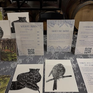 May include: A display of white paper prints featuring black ink illustrations of a snake and a bird, alongside informational signs with text including Wildlife Prints, About the Artist, and Custom Pet Commissions, all arranged on a lace tablecloth.