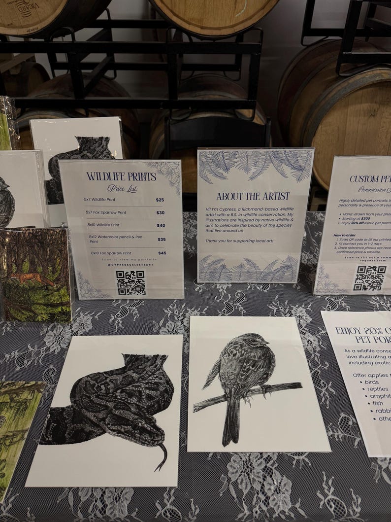 May include: A display of white paper prints featuring black ink illustrations of a snake and a bird, alongside informational signs with text including Wildlife Prints, About the Artist, and Custom Pet Commissions, all arranged on a lace tablecloth.