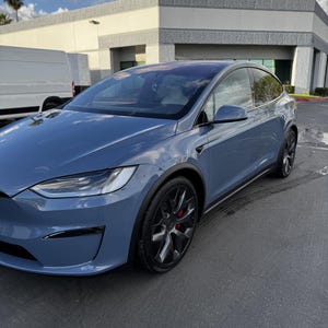 May include: A blue Tesla Model X electric SUV featuring a sleek aerodynamic body, dark multi-spoke alloy wheels, and visible red brake calipers. The vehicle has a panoramic windshield, black trim accents, and a modern, streamlined exterior design.