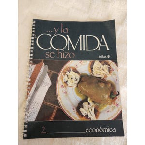 Könnte beinhalten: Ein spiralgebundenes Kochbuch mit dem Titel ...y la COMIDA se hizo mit schwarzem Einband, das ein Foto eines Tellers mit gefüllter Paprika und gezupftem Hähnchen, einer weißen Spitzenserviette und einem silbernen Messer zeigt.
