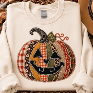 Puede incluir: Una sudadera de cuello redondo color crema con un gráfico central grande de una calabaza de Halloween. La calabaza presenta un diseño de patchwork con varios tejidos, incluyendo cuadros, flores y estampado de leopardo, con rasgos faciales negros cosidos.