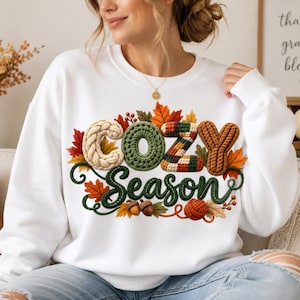 Puede incluir: Una sudadera blanca de cuello redondo con un diseño gráfico que incluye el texto "COZY Season" en una fuente texturizada similar al tejido. El diseño está rodeado de hojas de otoño coloridas, bellotas pequeñas y un ovillo de lana en tonos naranja, verde y marrón.
