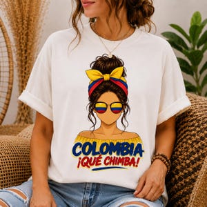 Puede incluir: Una camiseta blanca con el gráfico de una persona con un moño despeinado, usando una diadema y gafas de sol con los colores amarillo, azul y rojo de la bandera de Colombia. Debajo del gráfico, el texto dice COLOMBIA ¡QUÉ CHIMBA!
