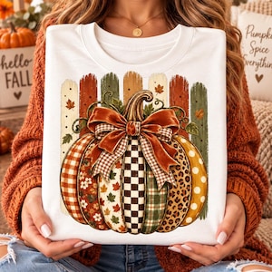 Puede incluir: Una sudadera blanca de cuello redondo con un gráfico de una calabaza con segmentos verticales de varios patrones, incluyendo cuadros, flores, ajedrez y estampado de leopardo. Un gran lazo naranja corona la calabaza frente a una valla de madera.