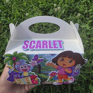 Puede incluir: Una caja de cartón blanca con asa que incluye una etiqueta personalizada con el nombre SCARLET en letras moradas. El frente de la caja muestra un gráfico colorido de Dora la Exploradora y el mono Botas en un entorno de selva con un mapa.