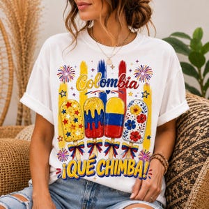 Puede incluir: Una camiseta blanca de manga corta con un gráfico de cuatro paletas heladas y el texto Colombia y Qué Chimba. El diseño incluye colores amarillo, azul y rojo, patrones florales, formas de estrellas y fuegos artificiales, con cada paleta atada con un lazo.