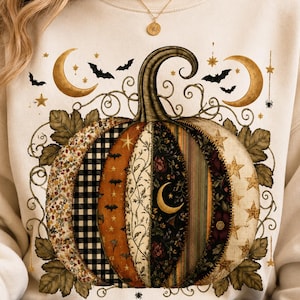 Puede incluir: Una sudadera de color crema con un gran gráfico central de calabaza compuesto por varios segmentos de tela estampada, incluyendo cuadros, flores y estrellas. El diseño incluye lunas crecientes, murciélagos y detalles de enredaderas.