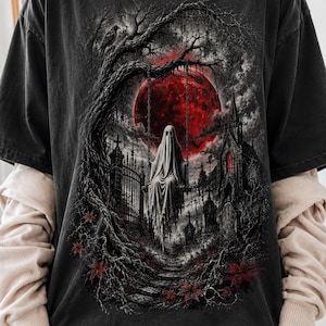 Puede incluir: Una camiseta gris carbón con un estampado gráfico de un fantasma sentado en un columpio suspendido de un árbol nudoso. La escena incluye una gran luna roja, un cementerio con lápidas, un edificio gótico y follaje oscuro con hojas rojas.