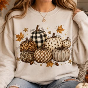 Puede incluir: Una sudadera de cuello redondo color crema con un gráfico central de varias calabazas con diversos patrones, incluyendo cuadros negros y blancos, estampado de leopardo, vichy marrón y lunares, decorado con estrellas doradas y hojas de otoño.