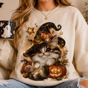 Puede incluir: Una sudadera beige de cuello redondo con un estampado gráfico de un gatito esponjoso con un sombrero de bruja negro, rodeado de calabazas naranjas, estrellas doradas y hojas de otoño. El gatito tiene ojos grandes y oscuros y bigotes largos.