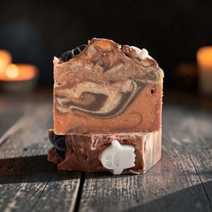 May include: A rectangular bar of soap featuring a marbled swirl of brown, tan, and orange tones. The soap is decorated with small, embedded black star-shaped accents and a white ghost-shaped figure on the front face of the bottom section.