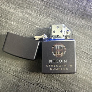 May include: A matte black metal flip-top lighter with a silver-toned insert. The front features a circular Bitcoin logo in gold and copper tones, with the text "BITCOIN" and "STRENGTH IN NUMBERS" printed in gold lettering below the graphic.