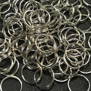 May include: A collection of silver-colored split key rings scattered on a dark surface. The key rings are round and made of metal, with a split design for easy attachment. The image shows a close-up view of the key rings, highlighting their shiny appearance.