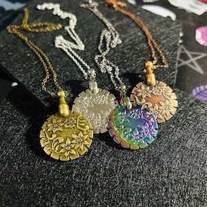 May include: Four floral pendant necklaces in gold, silver, iridescent, and copper tones. Each necklace features a round pendant with a floral design and a matching chain. The pendants are displayed on a black surface.