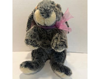 May include: A plush rabbit toy featuring mottled dark gray and black fur with a cream-colored muzzle and inner ears. The stuffed animal has long floppy ears and is adorned with a sheer, light pink ribbon tied in a bow around its neck.