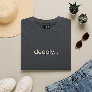 May include: A dark gray short-sleeved t-shirt featuring the word "deeply..." printed in a light beige font on the chest. The shirt includes a small black rectangular label at the inner neckline with the white text "HEK" printed on it.