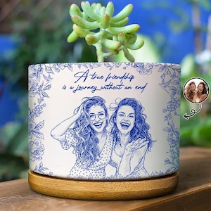 May include: A white cylindrical ceramic planter with a blue line art illustration of two smiling people and floral borders. The text reads, A true friendship is a journey without an end. The planter sits on a circular light wood base.