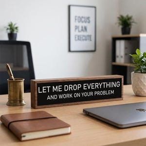 May include: A rectangular wooden desk sign with a black face and white text that reads, "LET ME DROP EVERYTHING AND WORK ON YOUR PROBLEM." The sign features a dark wood frame and is positioned on a desk surface alongside office supplies.