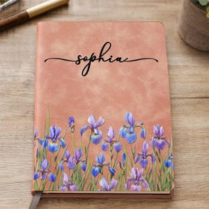 May include: A rectangular notebook with a textured peach-coloured cover featuring a printed illustration of purple iris flowers and green stems along the bottom. The name "sophia" is printed in black cursive script in the centre of the cover.