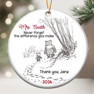 Puede incluir: Un adorno redondo de cerámica blanca con una ilustración en tinta negra de Winnie the Pooh y Piglet caminando. El texto dice: Mrs. Smith, Never forget the difference you make, Thank you, Jane, y el año 2026 en tinta roja.
