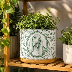 May include: A white ceramic planter featuring a green line-art illustration of a Basset Hound dog inside an ornate oval frame, surrounded by a botanical pattern of flowers and leaves, resting on a circular bamboo wood saucer base.