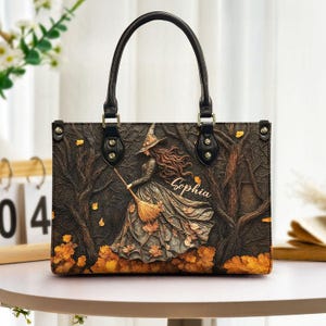 May include: A rectangular handbag with two black handles featuring a dark forest scene with a witch holding a broomstick. The design includes fallen orange leaves on the ground and the name Sophia written in a cursive script on the front panel.