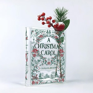 May include: A clear acrylic book-shaped vase featuring the printed title A Christmas Carol by Charles Dickens, decorated with green holly, red poinsettias, and top hats. A sprig of artificial pine needles and red berries is tucked inside the top.