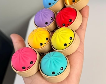 May include: A collection of eight small, round, colorful dumpling-shaped keychains, each resting in a miniature beige bamboo-style steamer basket. The dumplings feature smooth surfaces in blue, orange, purple, red, yellow, and pink, with printed black eyes and smiling mouths.