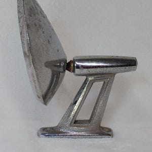 Peut inclure: Un rétroviseur latéral chromé vintage pour véhicule, doté d'une surface réfléchissante circulaire fixée à un bras de montage angulaire et effilé avec une base triangulaire creuse. La finition métallique présente des signes d'usure et de piqûres.