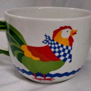 Peut inclure: Une tasse en céramique blanche ornée d'une illustration de coq coloré avec une crête rouge, une tête jaune et un corps vert. Le coq porte un foulard à carreaux bleus et blancs. La tasse possède une anse verte et une ligne ondulée bleue à la base.
