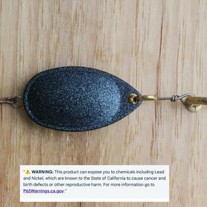 May include: A dark blue, glittery, oval-shaped fishing lure featuring a metallic treble hook on one end and a brass-coloured swivel ring on the other. A white rectangular warning label is present, stating: WARNING: This product can expose you to chemicals including Lead and Nickel, which are known to the State of California to cause cancer and birth defects or other reproductive harm. For more information go to P65Warnings.ca.gov.