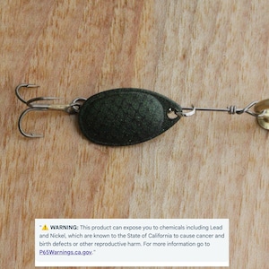 May include: A dark green oval fishing spoon lure with a textured scale pattern, featuring a silver-toned treble hook on one end and a gold-toned metal eyelet on the other. A warning label regarding lead and nickel exposure is visible at the bottom.