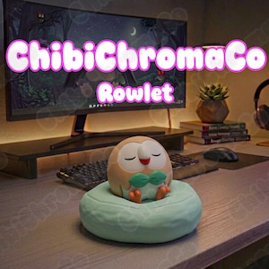 Puede incluir: Una figura redonda de color beige y blanco del personaje Rowlet con una pajarita verde, pies naranjas y ojos cerrados, sobre una base circular de color verde claro. El texto ChibiChromaCo Rowlet aparece en grandes letras blancas.