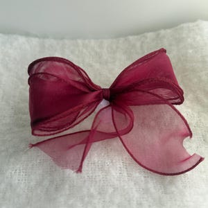 May include: A deep burgundy bow crafted from sheer, translucent organza ribbon. The bow features multiple layered loops and two trailing ends, showcasing a delicate, lightweight texture with visible edges and a consistent dark red colour throughout.
