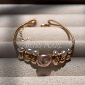 May include: A gold-toned wire-wrapped bracelet featuring a central large, irregular, iridescent pinkish-white pearl flanked by smaller white pearls. The metal wire is intricately woven around the pearls. The text BLISS PEARL is visible.